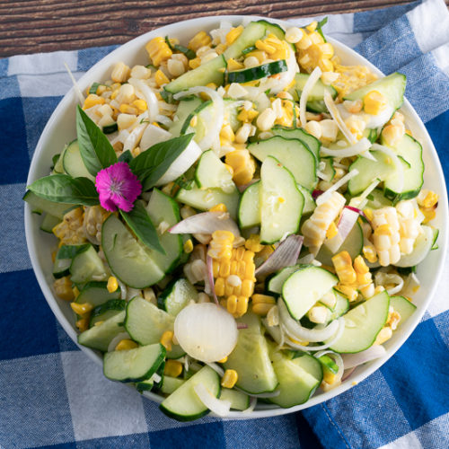 top down of cucumber and corn salad in a white bowl, on a blue checker napkin.