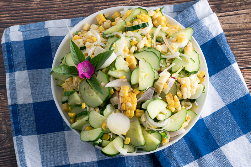 top down of cucumber and corn salad in a white bowl, on a blue checker napkin.
