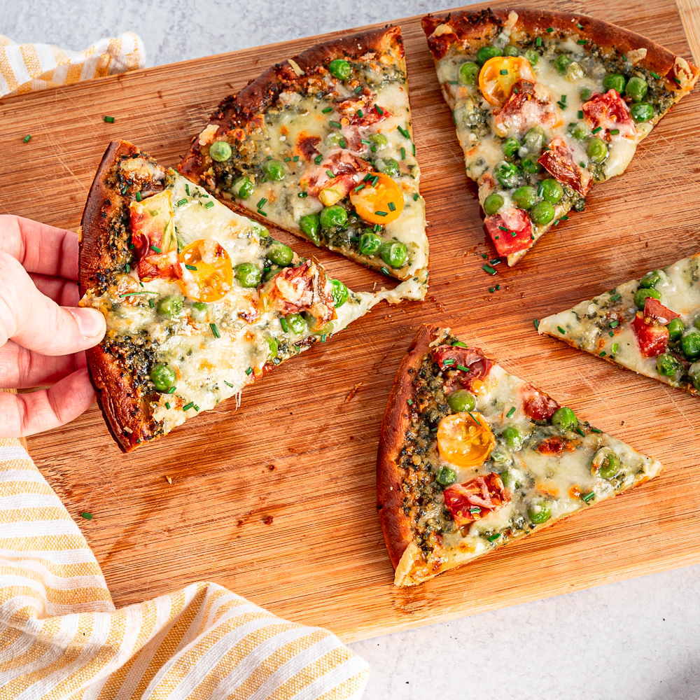 Pizza slices on a cutting board. With toppings like peas, pesto, and peppers. A hand is taking one slice off the board.