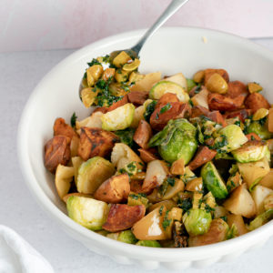 White bowl of Roasted Sweet Potatoes, Apples, and Brussel Sprouts with a silver spoon, spoon over an herbed chestnut topping.