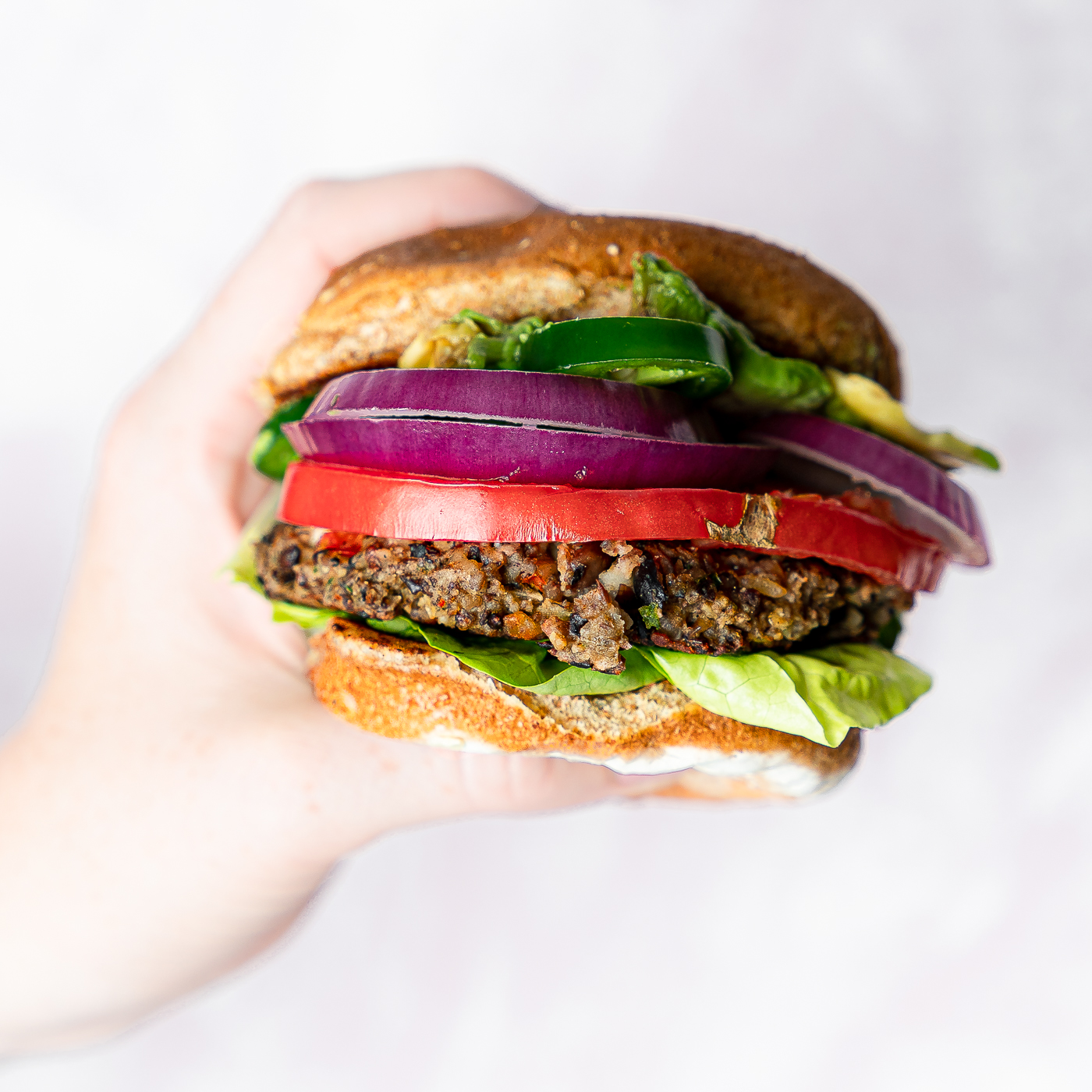 A hand holding a veggie burger with lettuce, red onion slices, a tomato slice, and fresh slices jalapeno. 