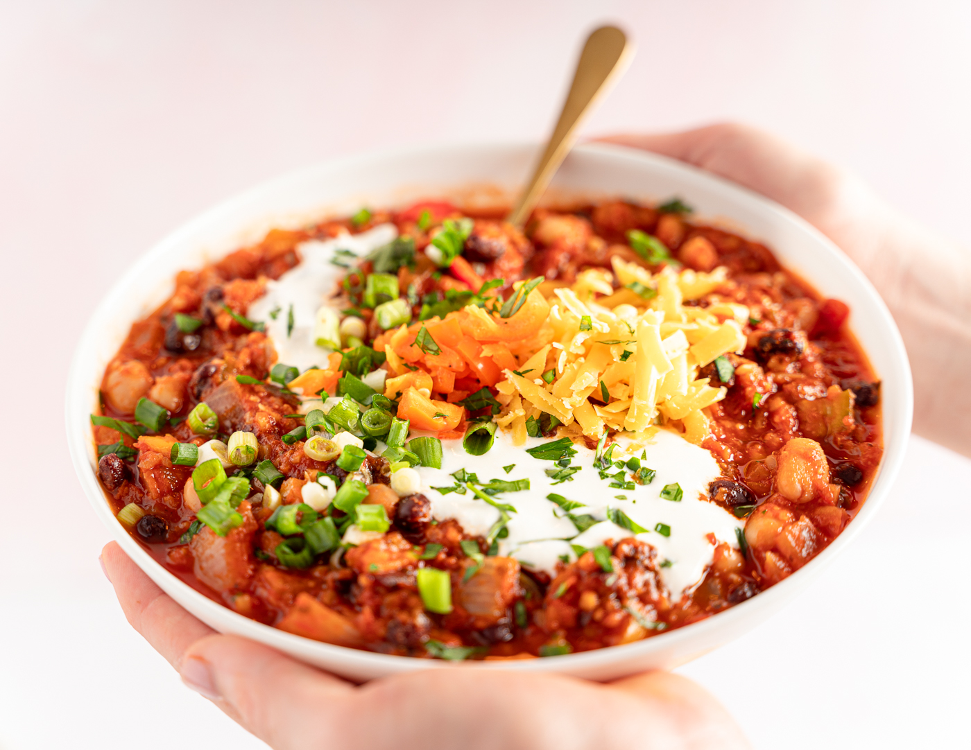 A bowl of Vegetable chili enclosed by hands. The chili is garnished with green onion, cheddar cheese, diced orange bell pepper, and creamy sour cream.