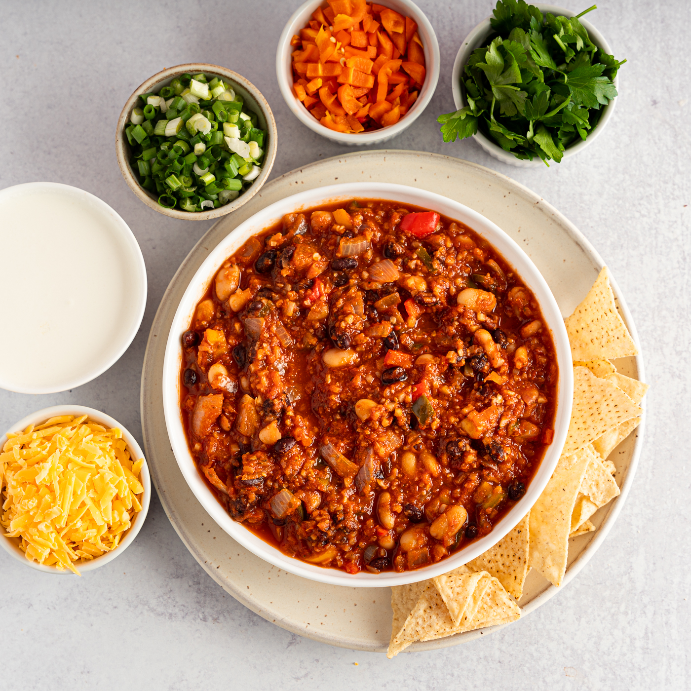 A bowl of vegetable chili on a plate with tortilla chips. Around the bowl are smaller bowls full of garnishes such as cheddar cheese, sour cream, green onion, orange bell pepper, and herbs. 