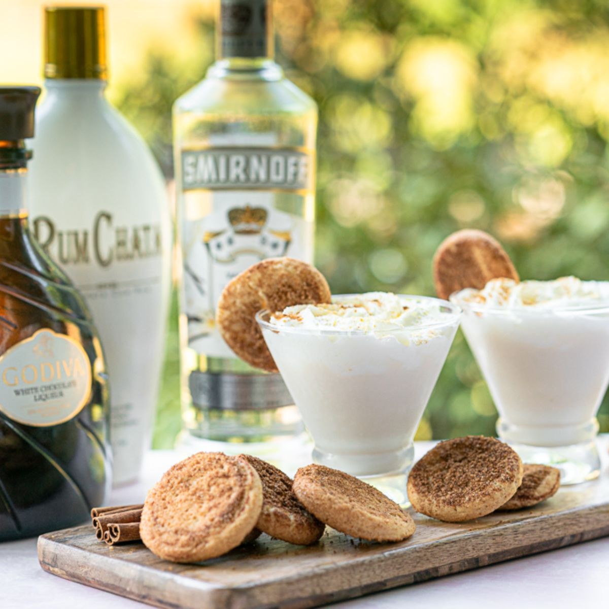 two snickerdoodle martinis with snickerdoodle cookies as garnish. The ingredients in the background, vanilla vodka, rumchata, and white chocolate liquor. 