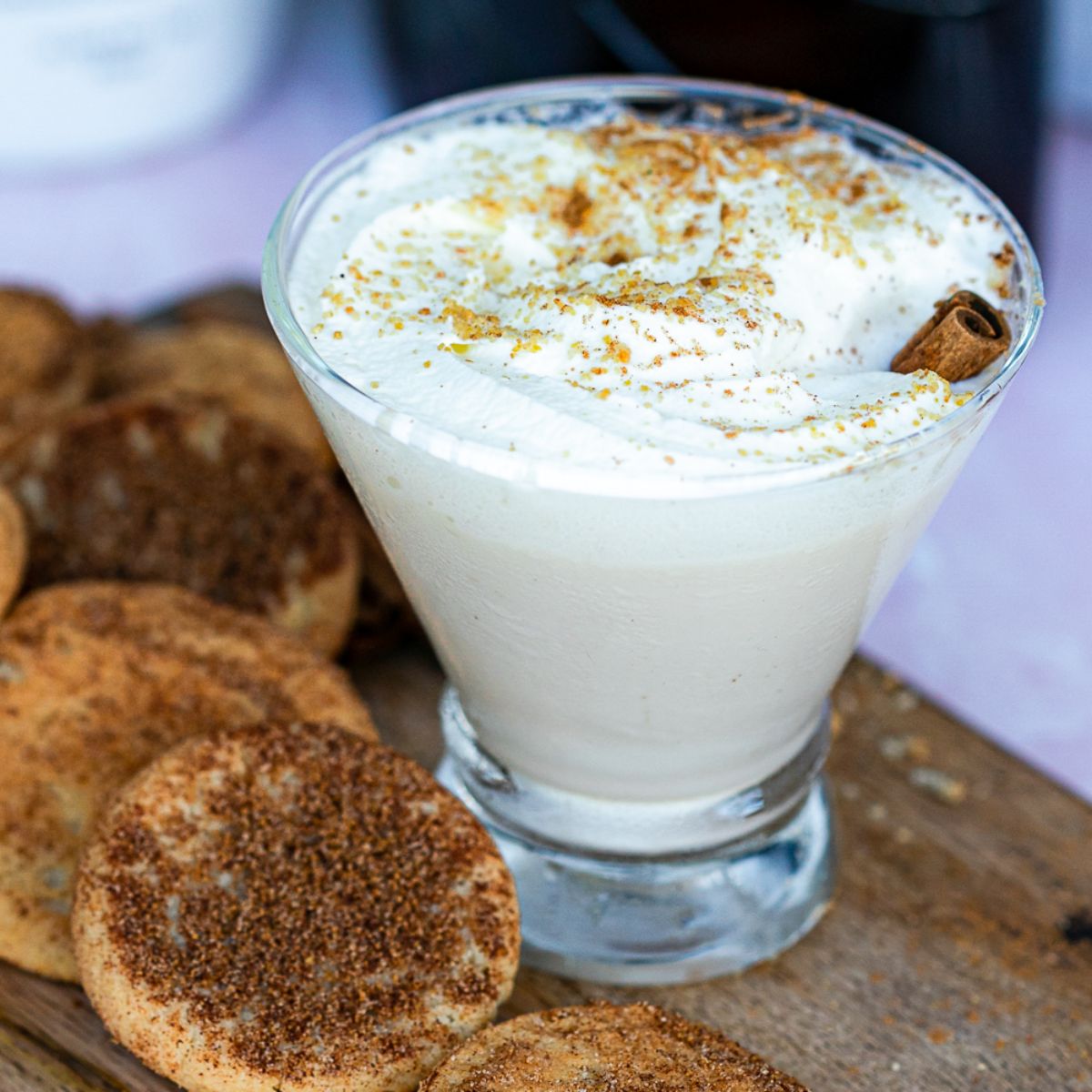 snickerdoodle martini with a creamy foam topping, a cinnamon stick garnish, and snickerdoodle cookies surrounding the cocktail. 