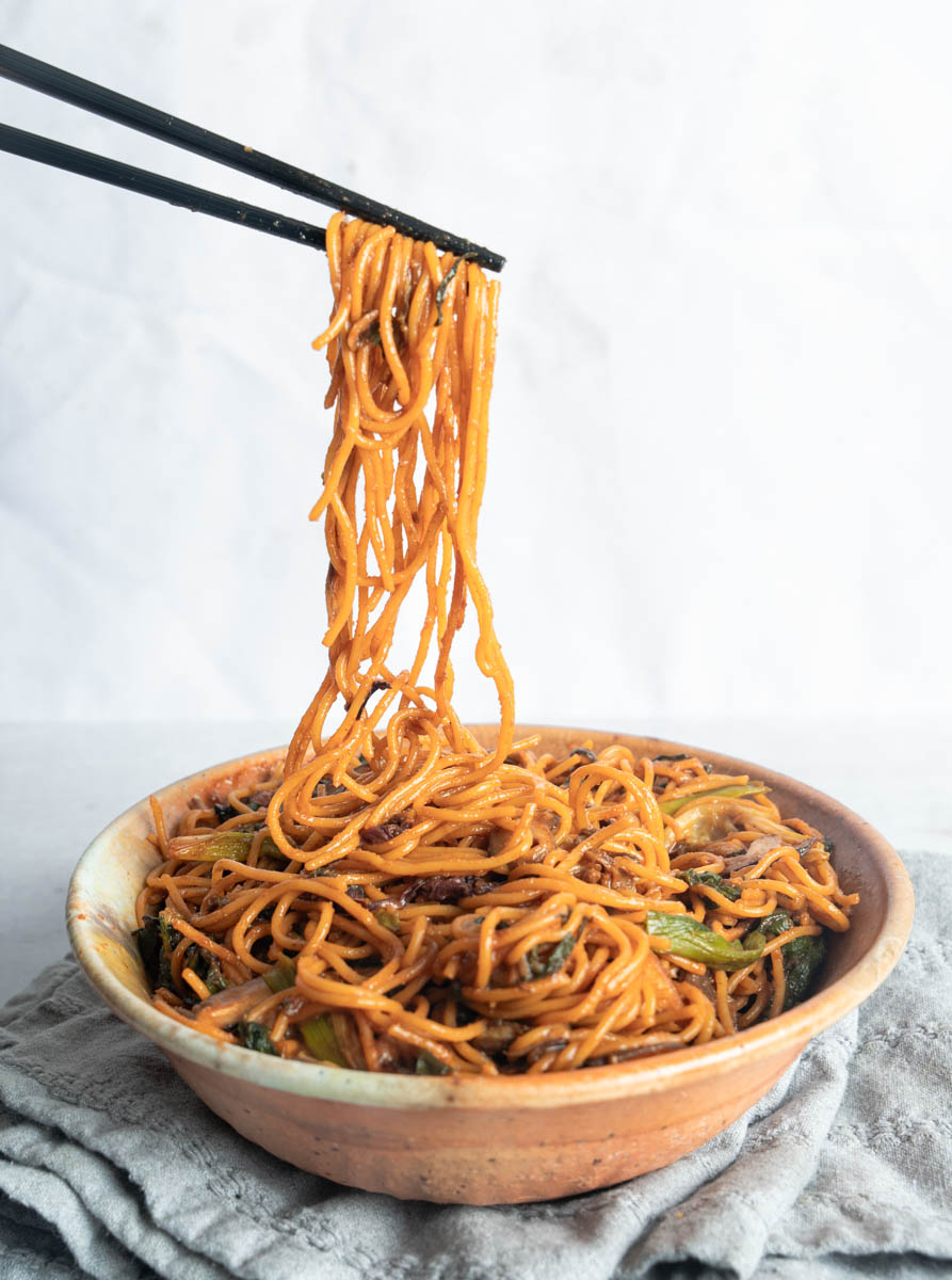 Vegan Yakisoba noodles in a bowl over top a grey napkin. Black chopsticks are pulling noodles high above the brown bowl full of noodles.