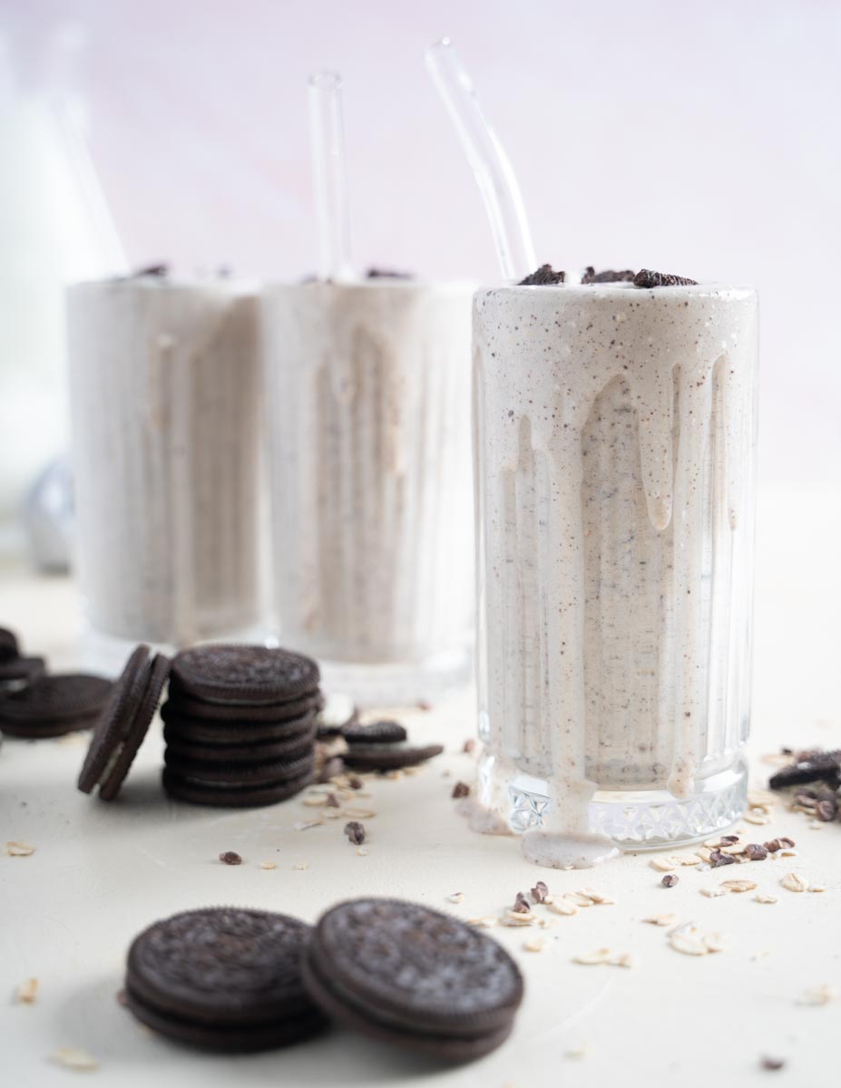 Three oreo milkshakes without ice cream, tow of which are in the back and out of focus, one is off center and in focus with milkshake dripping down the side and crushed oreos sprinkled over top.