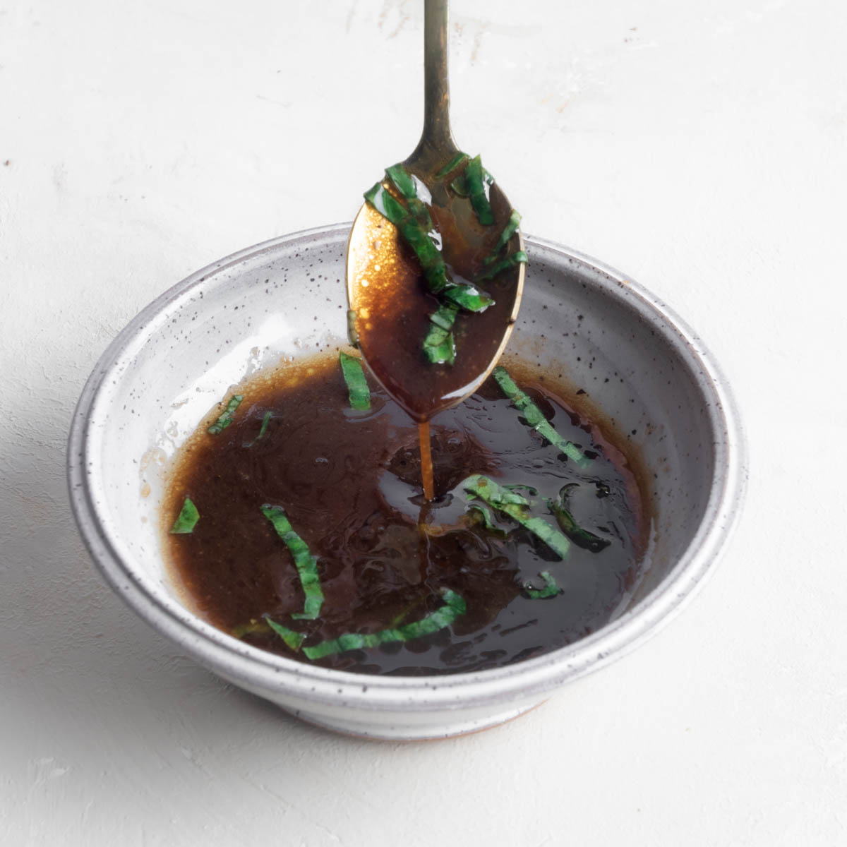 Basil balsamic vinaigrette in a white bowl with a spoon hovering over the deep brown dressing dripping the vinaigrette back into the bowl with julienned basil mixed throughout