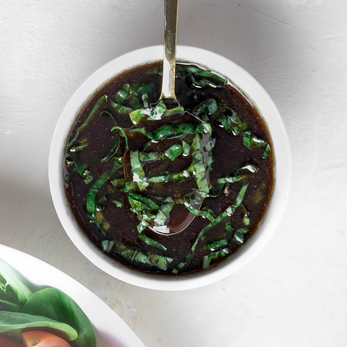 Basil balsamic vinaigrette in a white bowl with a spoon hovering over the deep brown dressing with julienned basil mixed throughout