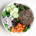 A large black bean burger patty laid overtop a spinach salad with cucumbers, shallots, and sliced cherry tomatoes.