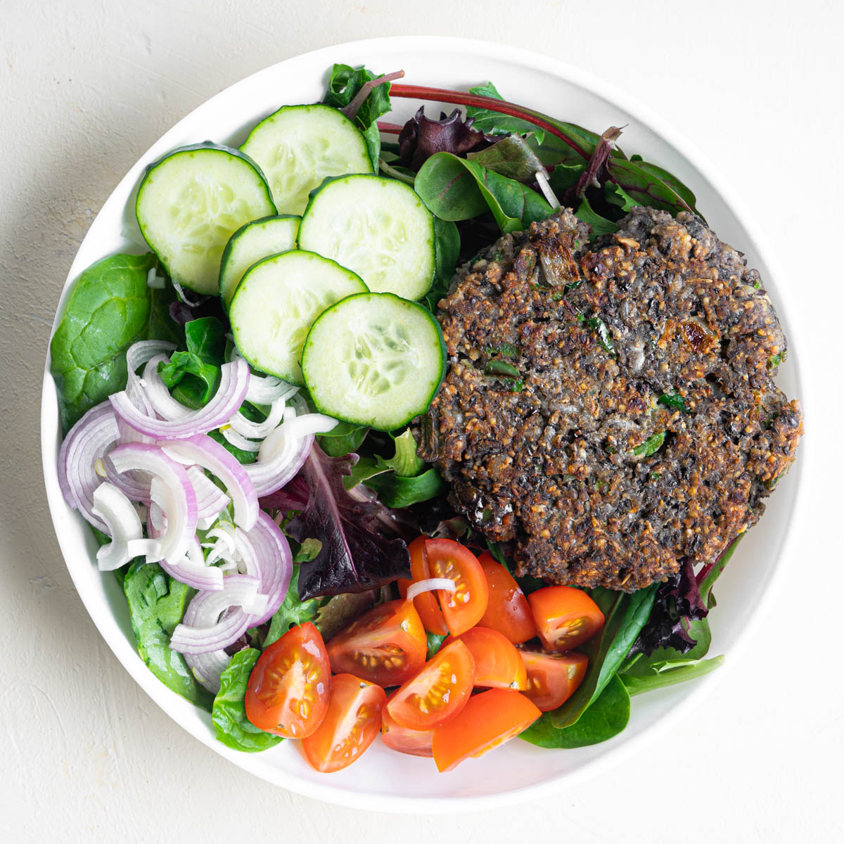 A large black bean burger patty laid overtop a spinach salad with cucumbers, shallots, and sliced cherry tomatoes.