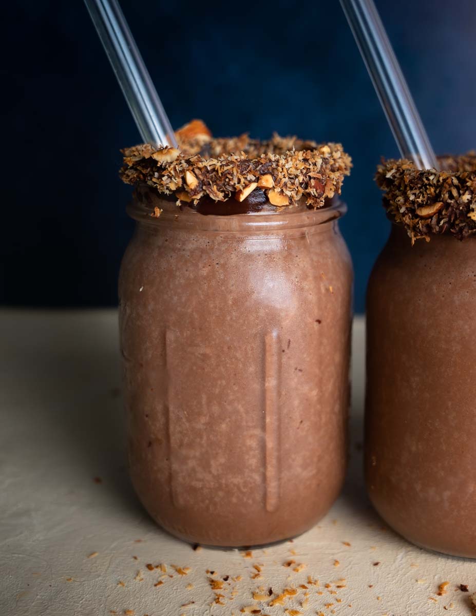 A chocolate brown smoothies in a mason jar rimmed with chocolate, toasted coconut, and chopped almonds
