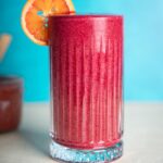 Berry_Rhubarb_Smoothie in a glass with a sliced orange garnish. Blue background.