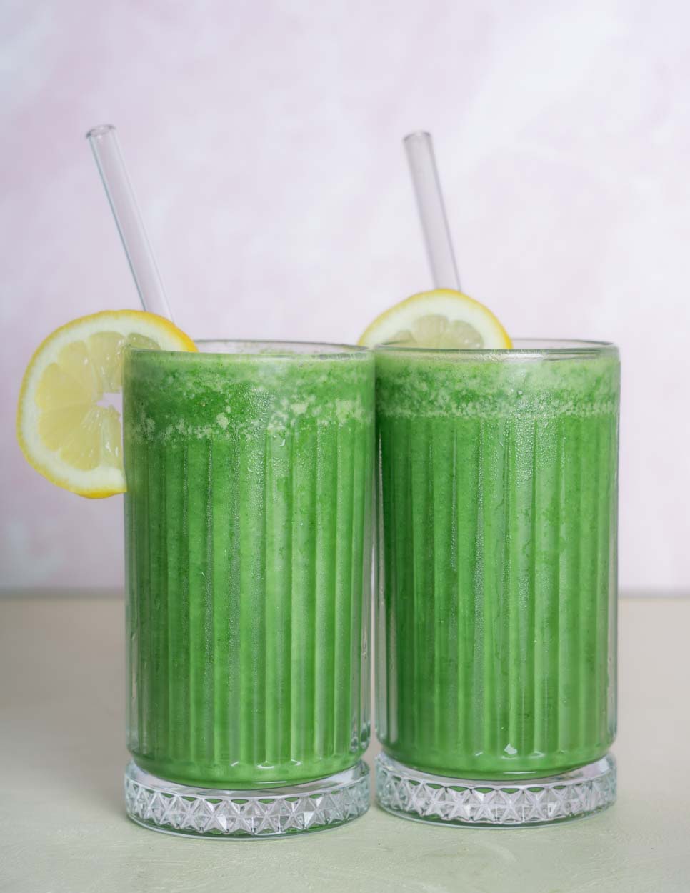 Two glasses of green juice smoothies with a slice of lemon as a garnish. Set in front of a light pink background