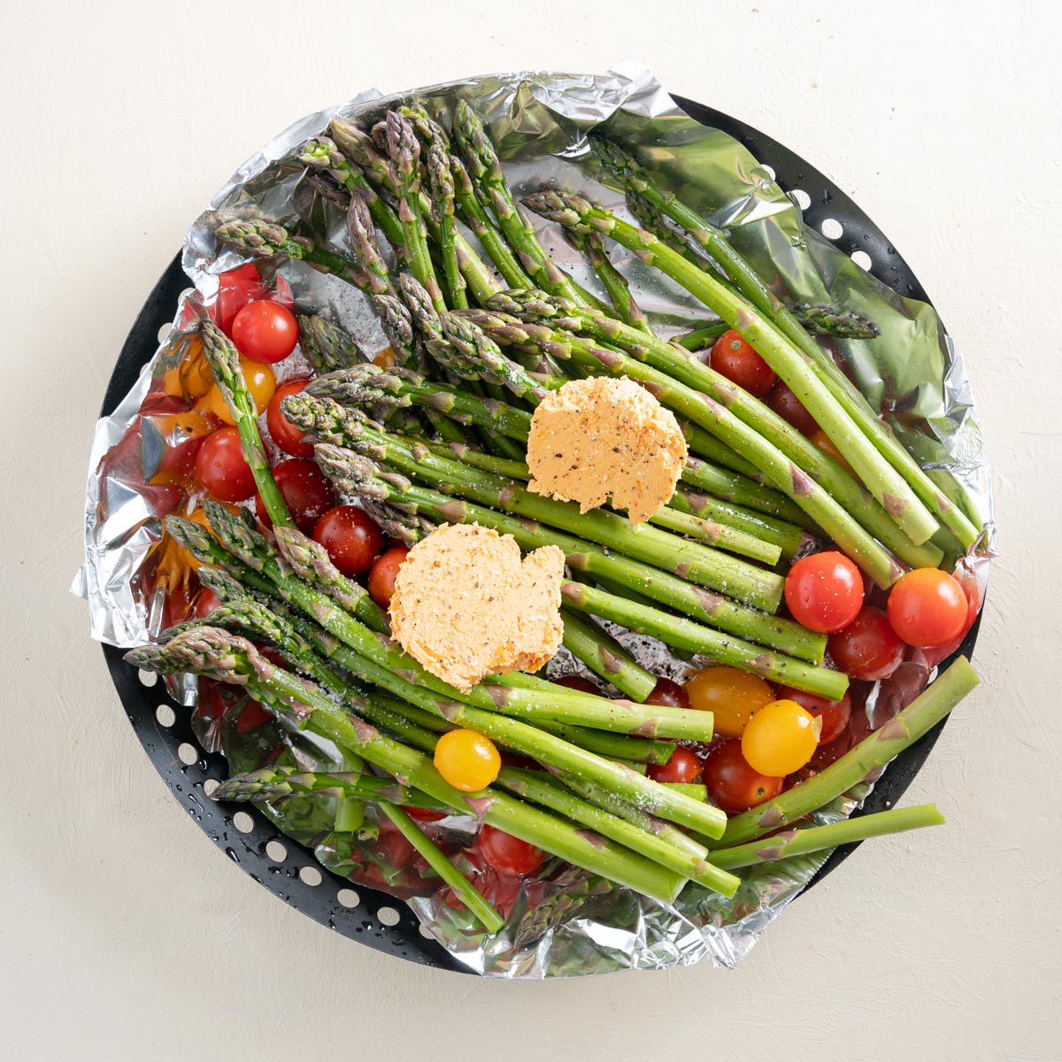 Raw asparagus and tomatoes in a grill pan with 2 large pats of tomato butter over top