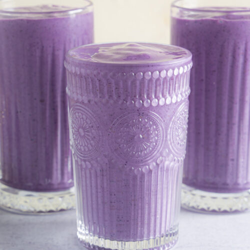 Berry Bliss Smoothie, a close up of three glasses with purple-blue smoothie inside. The glass is a unique design compared to the other two glasses. The photo is straight on and slightly elevated.