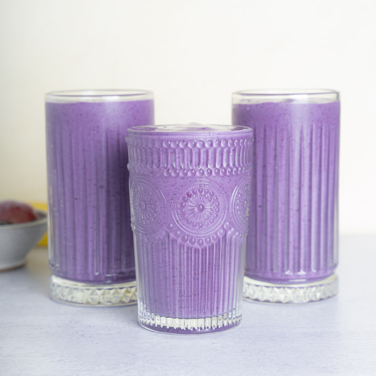 Three glasses of purple-blue colored beverage, the Berry Bliss Smoothie. The photo is take straight on with a small bowl of berries on the right slide of the frame. The background is cream.