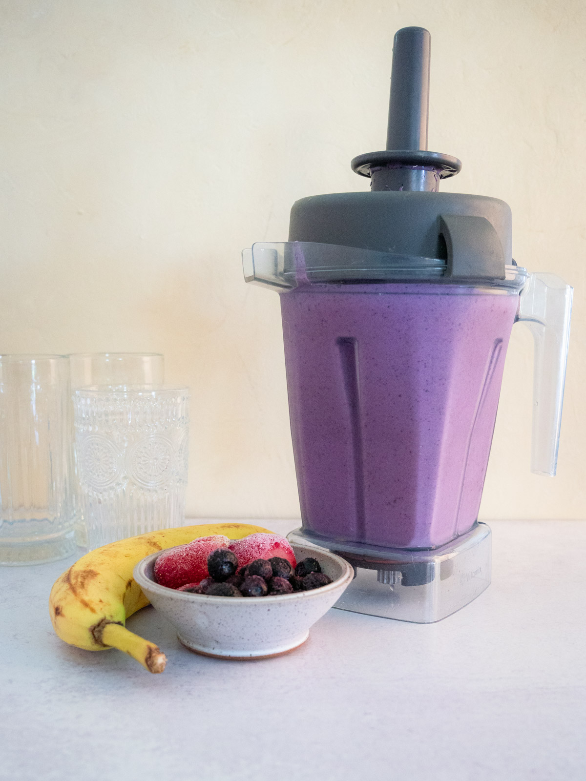 Berry Bliss Smoothie is in a vitamix blender with glasses, one ripe banana, and frozen berries in the scene. The shot is strait on with a cream back drop.