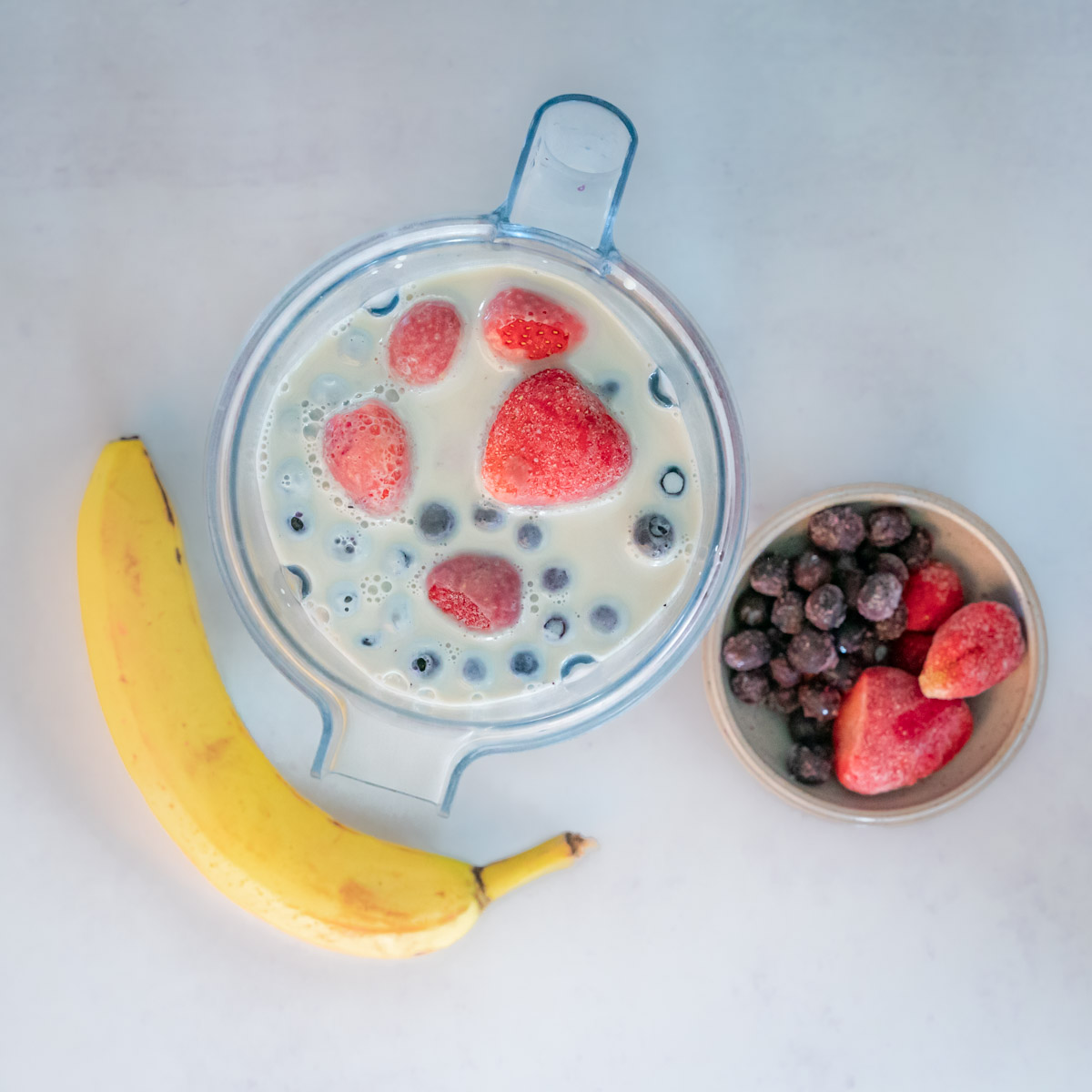 Top down of smoothie ingredients. In the photo include banana, a blender container with milk and berries visible, and a small bowl to the right of the blender full of berries.