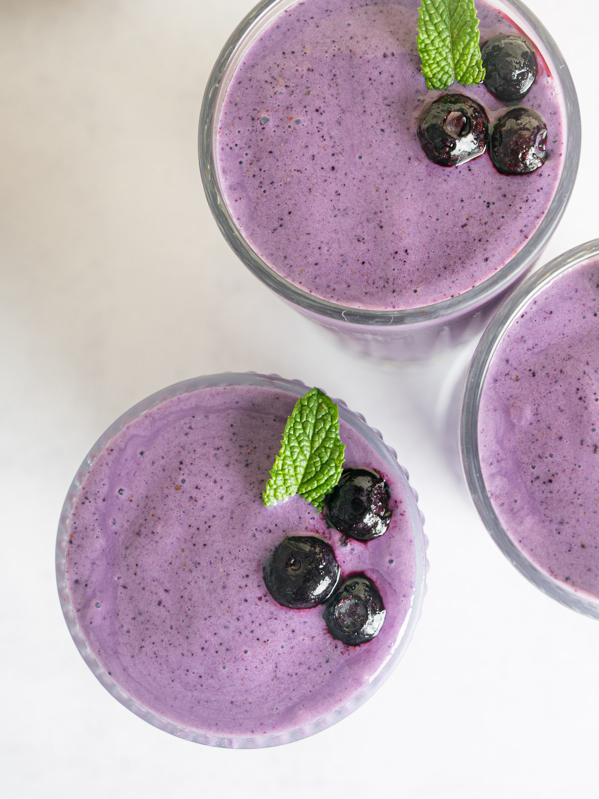 Top down of the Berry Bliss Smoothie. Three glasses with garnish over top. The garnish includes three blueberries and a mint leaf.
