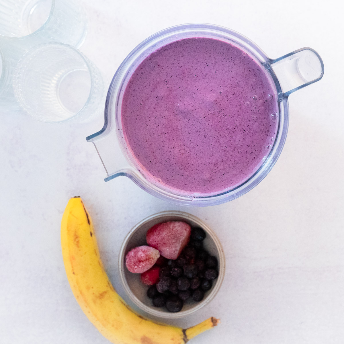 Top down Berry Blast smoothie in blender with ingredients in the shot. Seen is one ripe banana and a small bowl with berries. Decorative glasses are on the top left corner and the entire seen is on a cream background. 