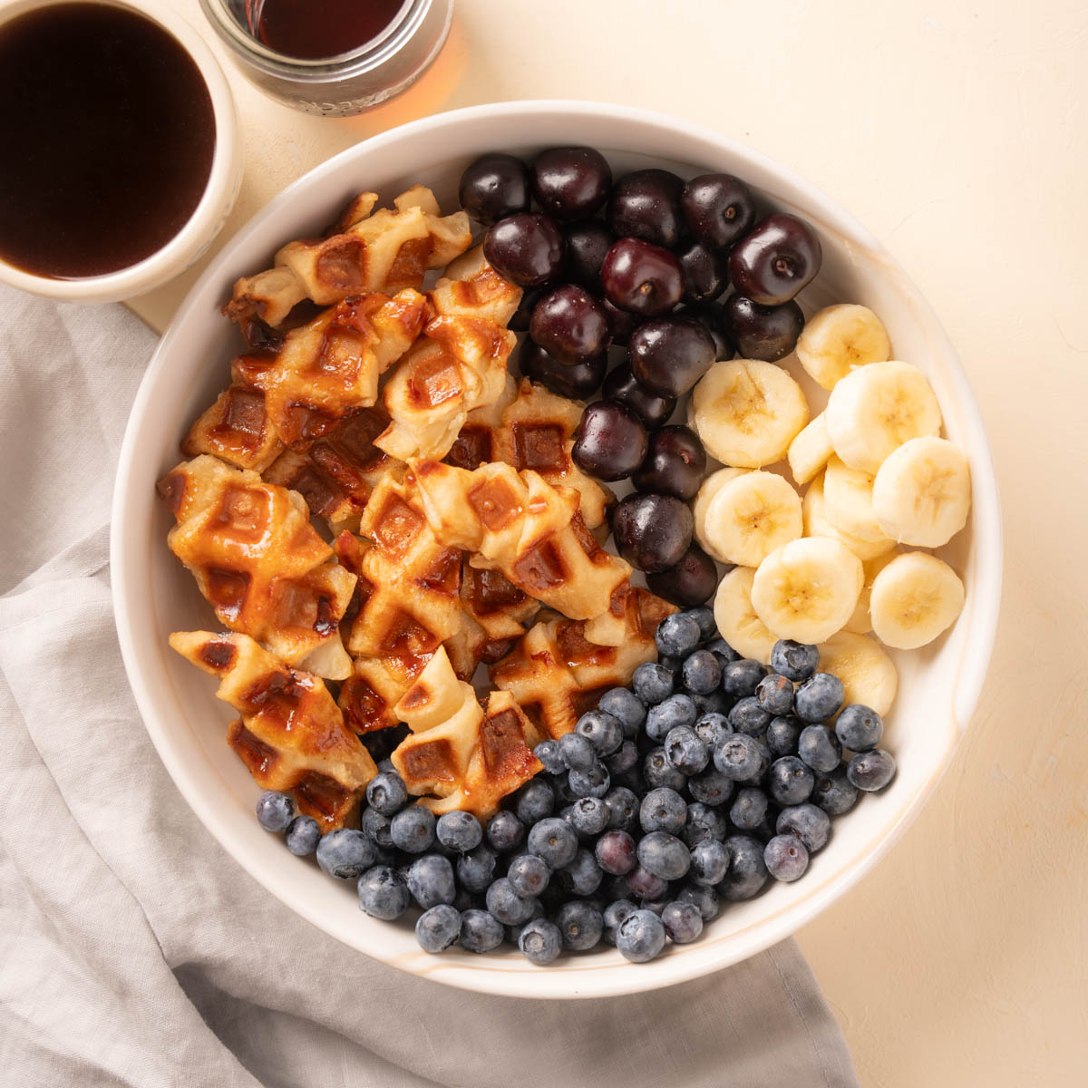 Large bowl of croffles (croissant waffles) with blueberryies, cherries, and sliced banana. On the side there is coffee and syrup. 