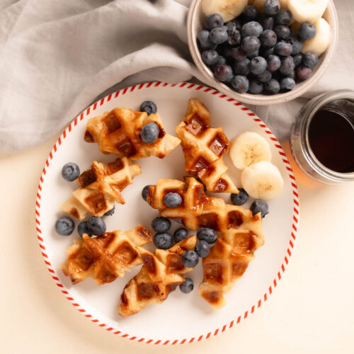 A plate with 7 croffles (croissant waffles). Blueberries sprinkled over top with a side of blueberries, sliced banana, and syrup.
