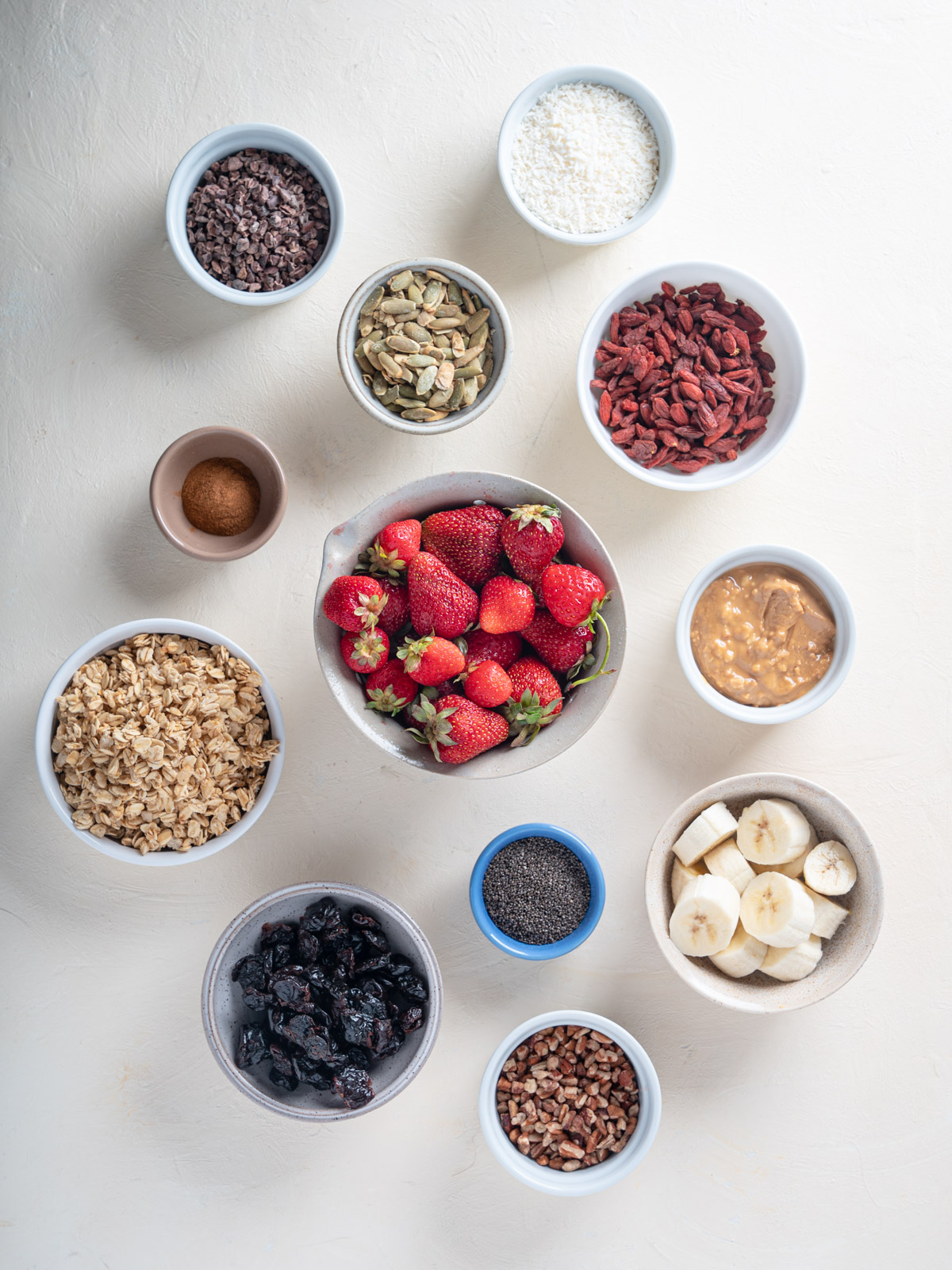Top down of ingredients in various bowls. The ingredients include whole red strawberries, sliced banana, granola, goji berries, chia seeds, pecan pieces, coconut flakes, cinnamon, peanut butter, pumpkin seeds, and cocoa nibs. Bowls are placed on a cream background.