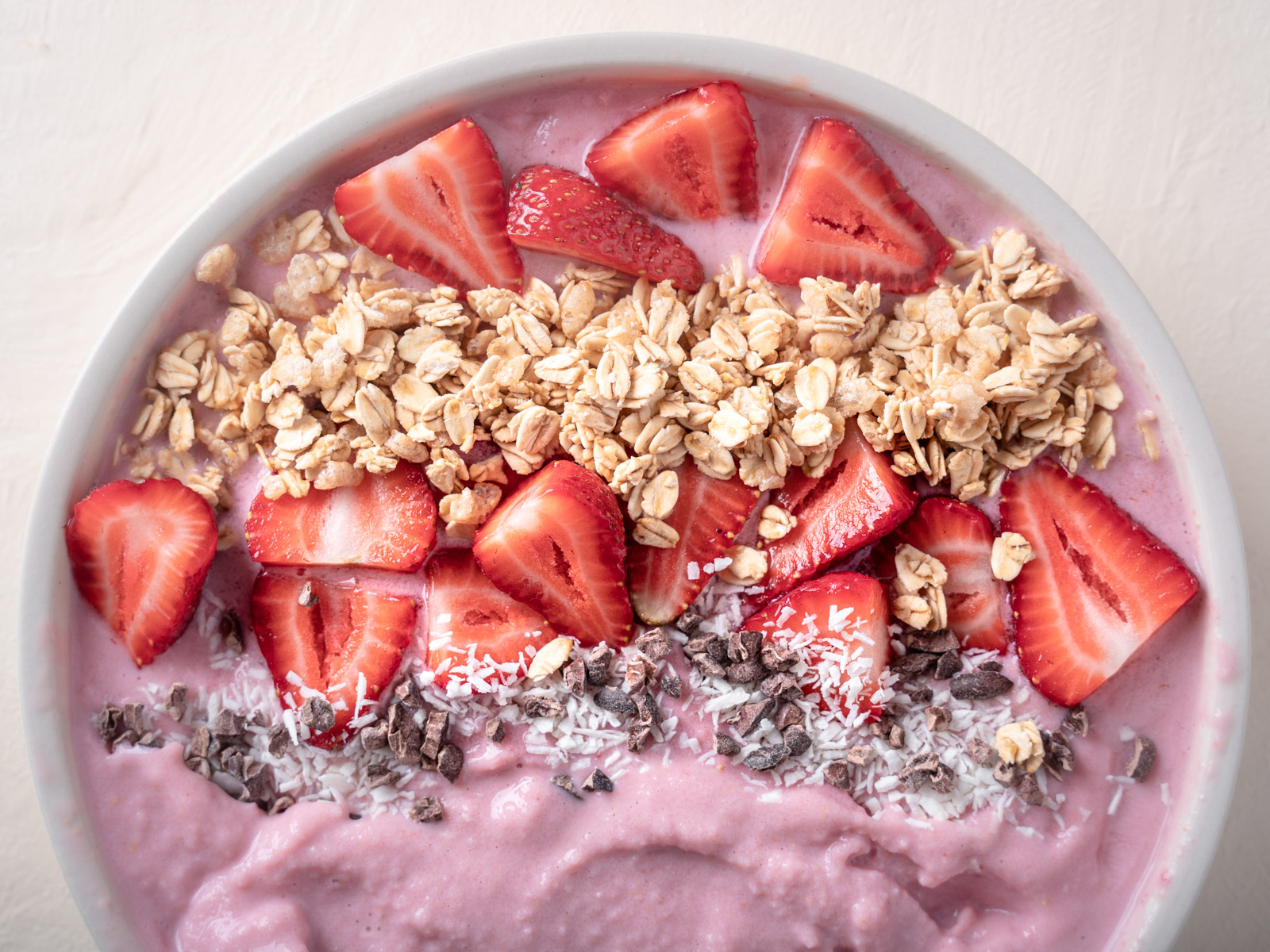 Close up of the strawberry smoothie bowl with toppings. The smoothie base is light pink and the toppings include cocoa nibs, coconut flakes, sliced strawberries, and granola.