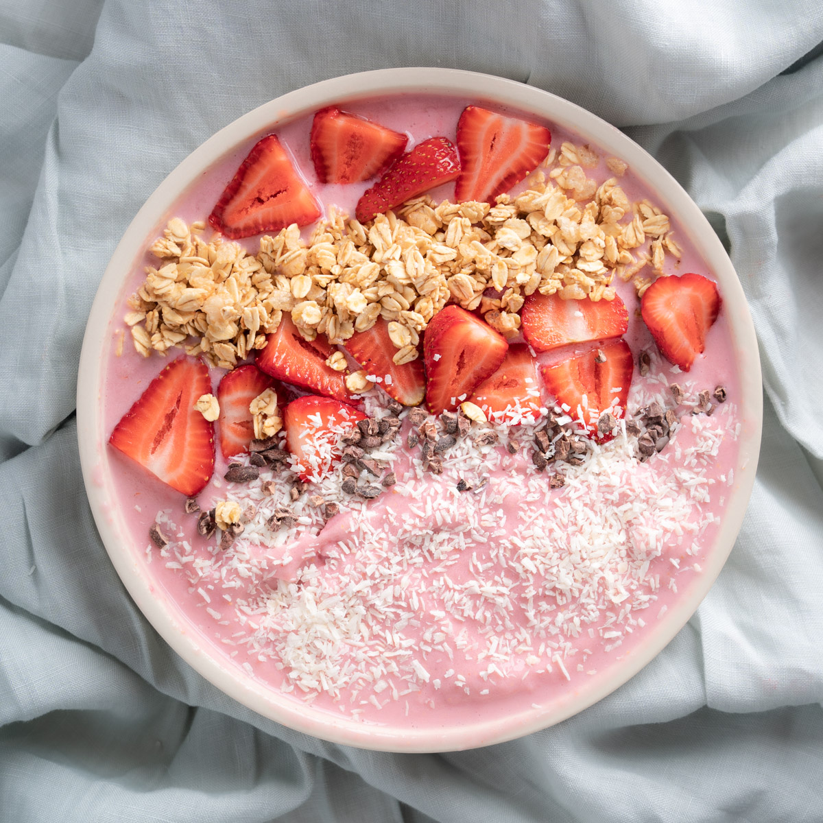 Strawberry Smoothie Bowl with pink smoothie base. Smoothie bowl is topped with coconut flakes, sliced strawberries, granola, and cocoa nibs. Bowl is placed on a sage linen table cloth. 