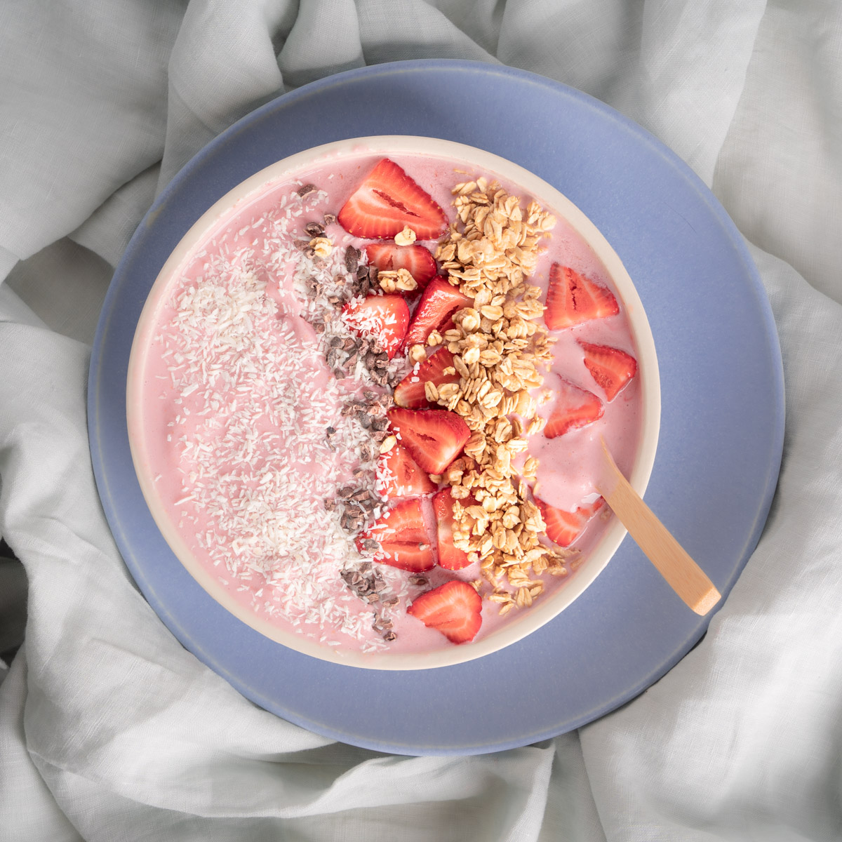 Strawberry Smoothie Bowl. Pink colored smoothie base with coconut flake, cocoa nib, sliced strawberry, and granola toppings. Placed on a blue plate over a sage linen table cloth.