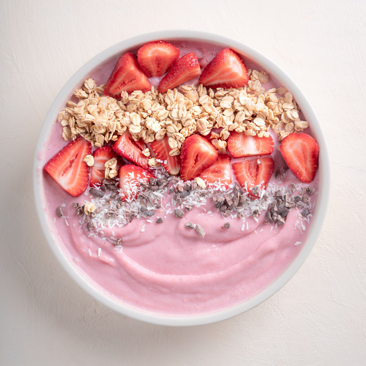 Strawberry Smoothie Bowl with toppings such as granola , sliced strawberries, coconut flakes, and cocoa nibs. Placed on a plain eggshell white back drop.