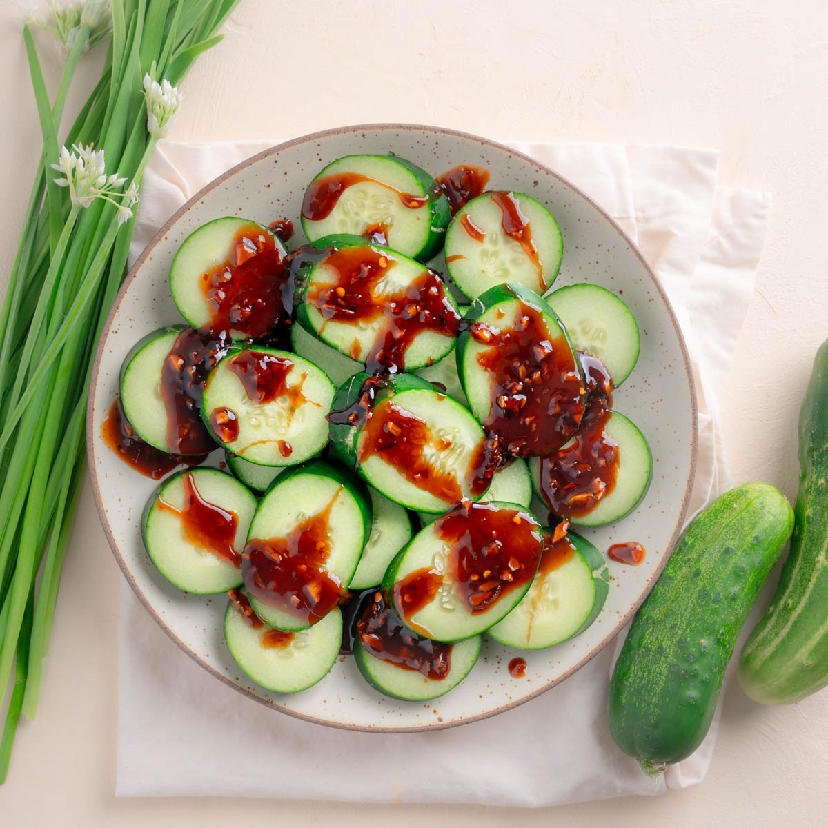 Top down of sliced cucumbers with a Garlic Sweet Soy Glaze Topping on a speckled plate. Cucumbers to the right as a prop and Chinese Garlic Leeks to the left with white flowers. 