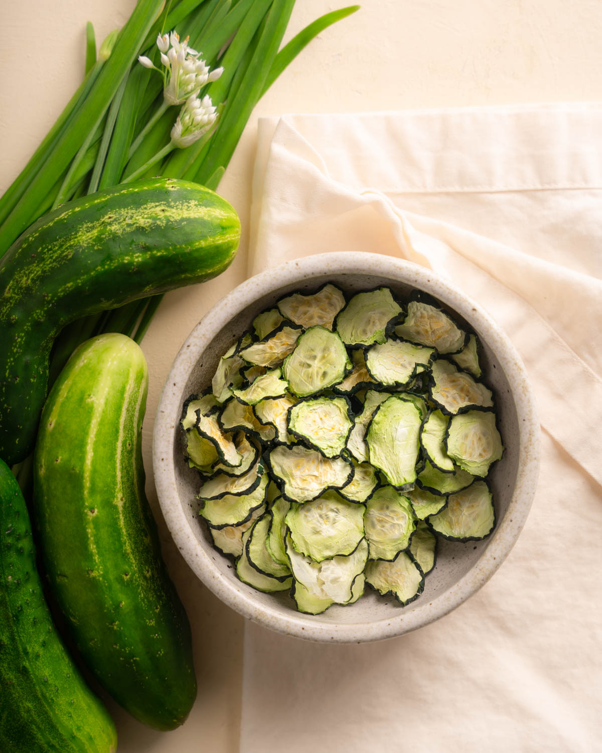 Dehydrated cucumbers (cucumber chips) in a bowl. There are cucumber and chionese garlic leek with white flower tops as props to the left side of the bowl. 