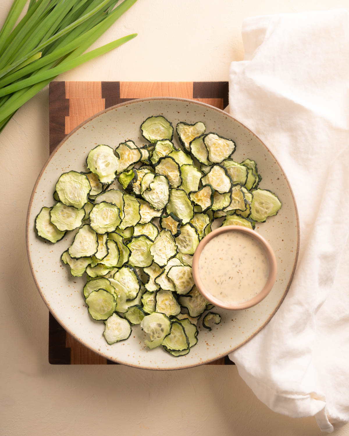 A plate of dehydrate cucumbers (cucumber chips) surround with a linin napkin, a bowl of ranch, and some green edible prop to the side. The plate is sitting on top of a two toned board.