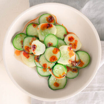 Cucumbers sliced and stacked in an off white bowl. The cucumber salad has a glistening vinegar sauce with a drizzle of red chili oil over top.