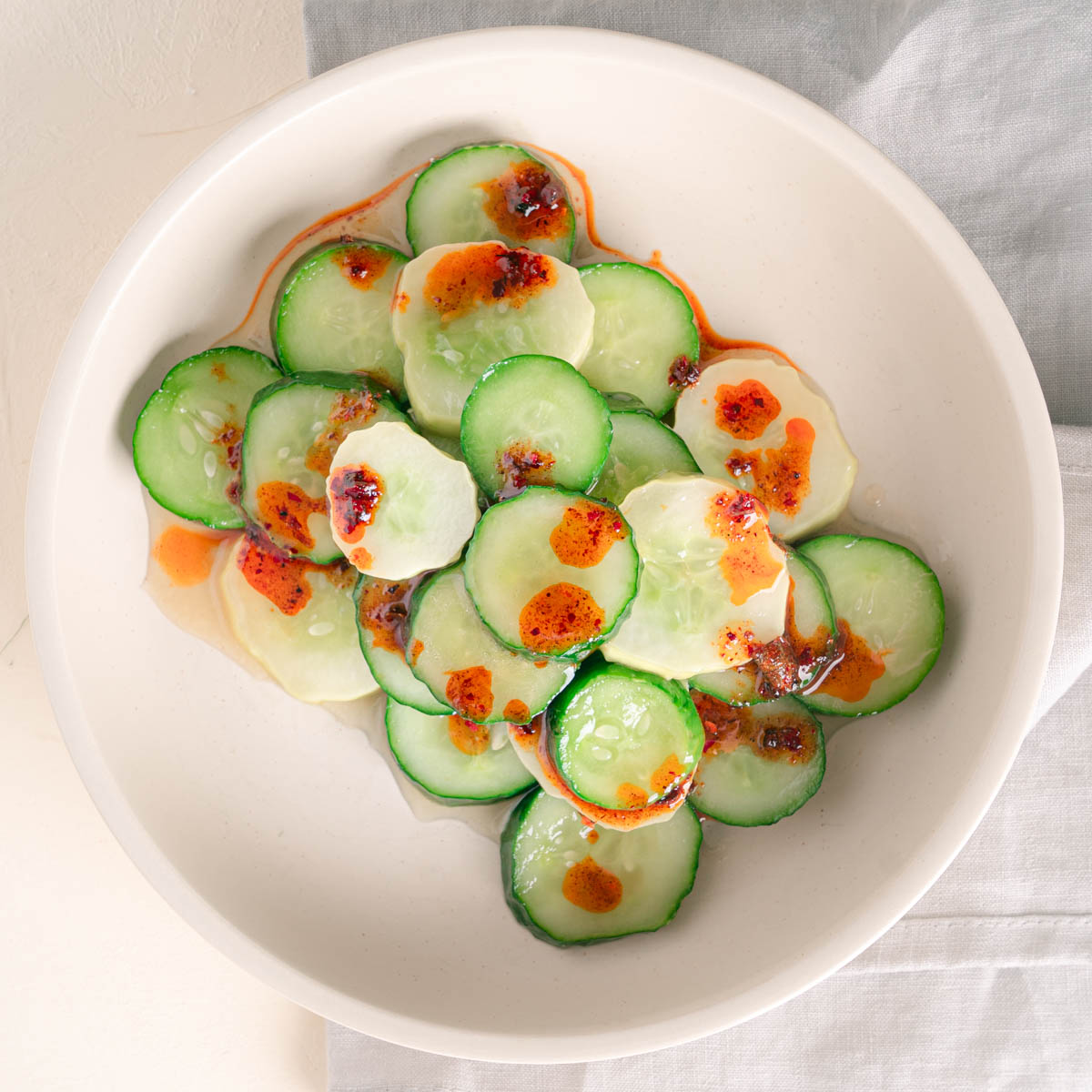 Cucumbers sliced and stacked in an off white bowl. The cucumber salad has a glistening vinegar sauce with a drizzle of red chili oil over top.