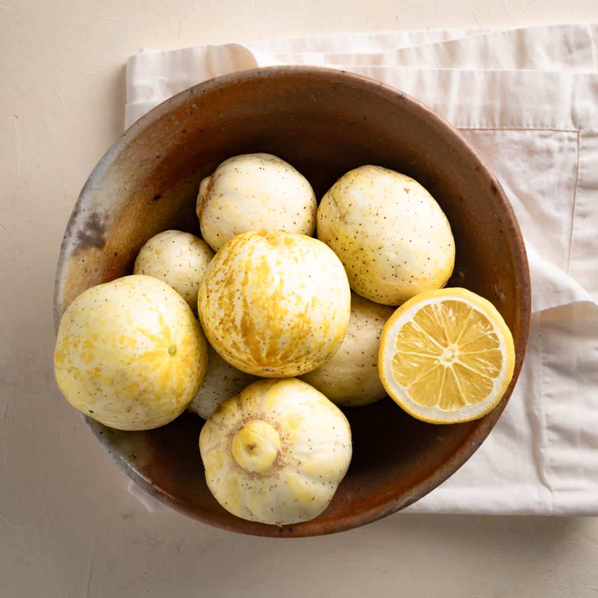 Top down view of lemon cucumbers in a brown bowl atop an off white linen napkin. Half a lemon is sitting cut side up in the bowl.
