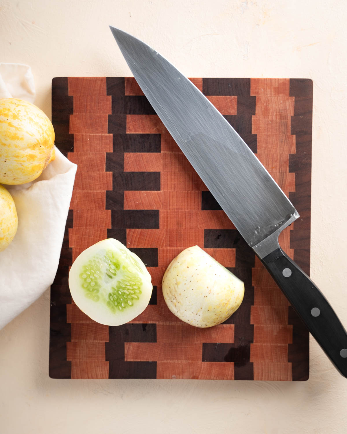 A lemon cucumber sliced in half on a wooden cutting board. The knife is in the shot and some unsliced lemon cucumbers are off to the left. 