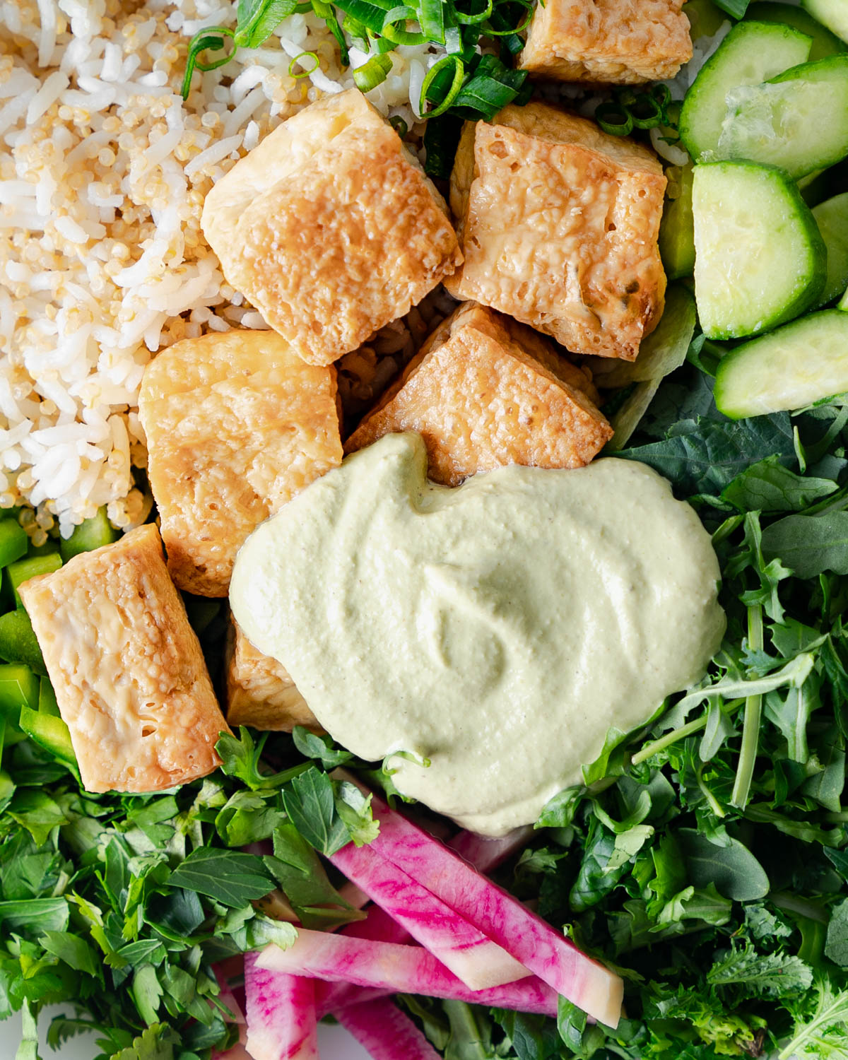 close up of baked tofu, rice and quinoa, cucumbers, chopped herbs, green pepper, watermelon rasdish sticks, and salad greens in a bowl as a salad. Topped with Creamy cashew dressing.
