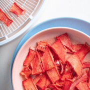 Top down view of dehydrated watermelon on a pink plate over top a larger blue plate. A dehydrator tray with dehydrated watermelon is above the plate to the left.