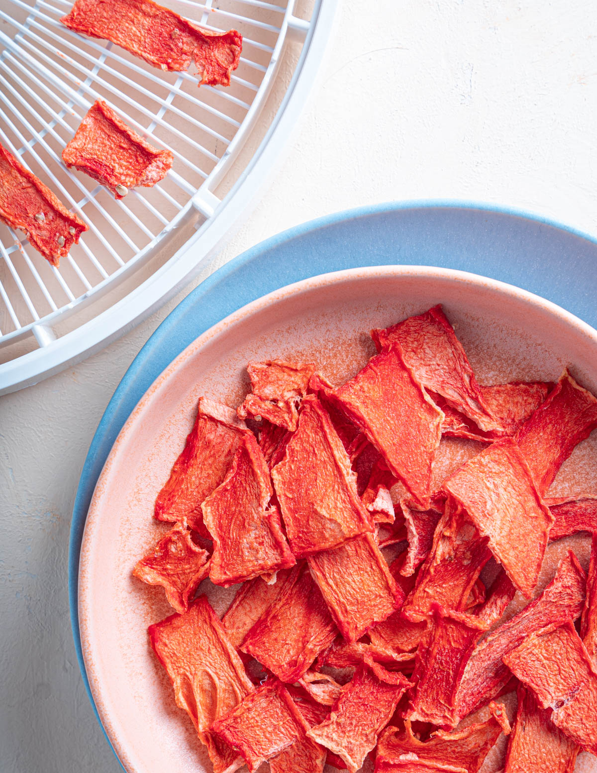 Top down view of dehydrated watermelon on a pink plate over top a larger blue plate. A dehydrator tray with dehydrated watermelon is above the plate to the left.