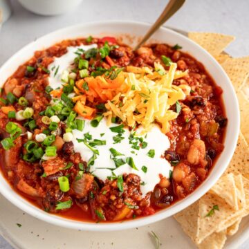 Vegan Vegetable Chili with tortilla chips to the right of the bowl. Toppings include vegan sour cream, cheese, orange bell peppers, and green onions.