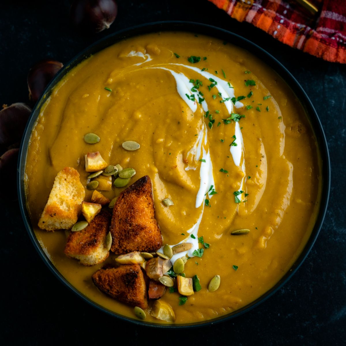 golden orange pumpkin soup garnished with pumpkin seeds, a cream sauce, golden brown croutons, and fresh herbs.