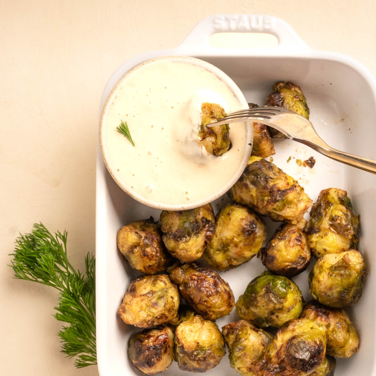 A stoneware bowl with a creamy Horseradish Aioli inside a casserole dish with air fried brussel sprouts. A fork is dipping one Brussel Sprout into the dip.