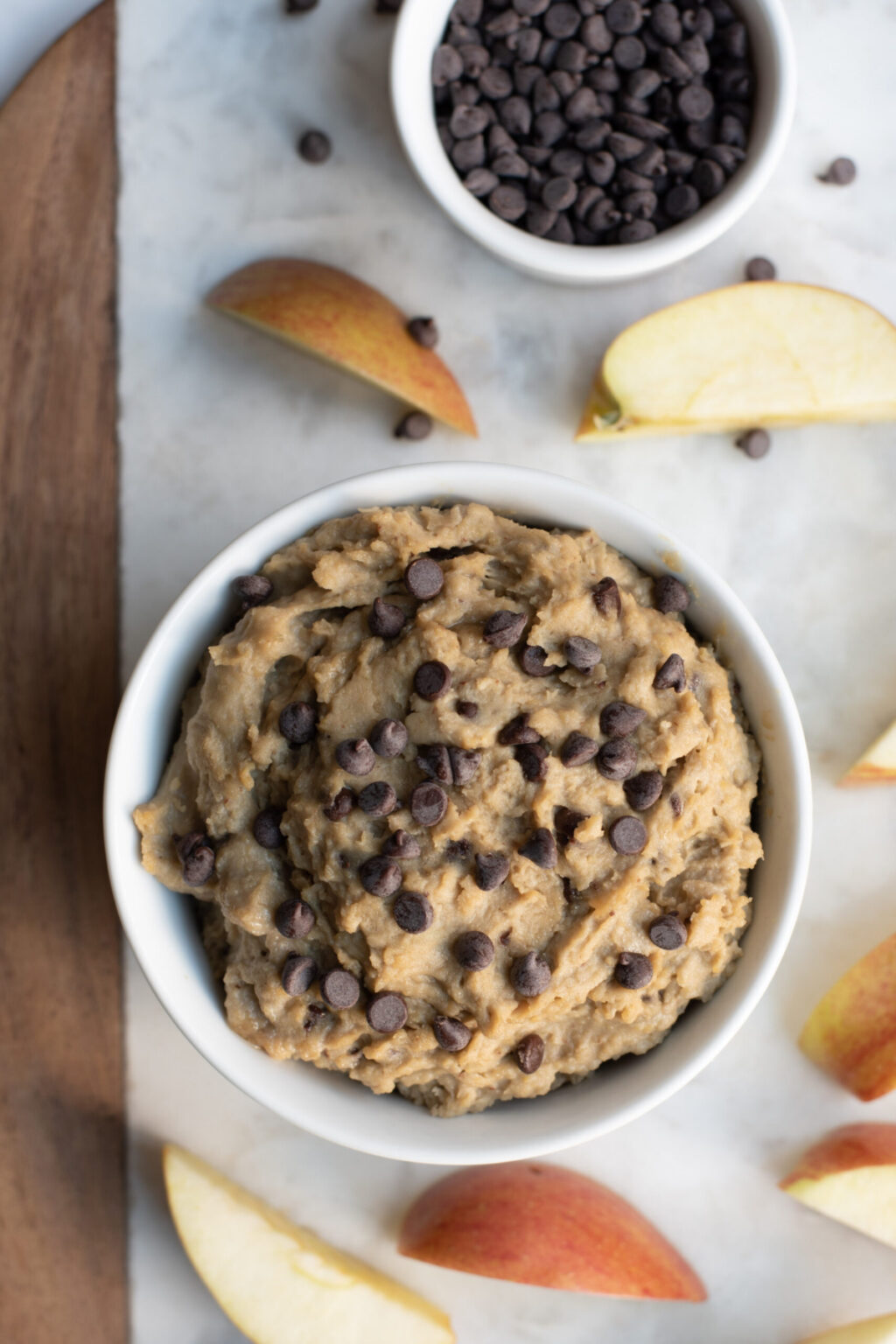 Chickpea Cookie Dough Dip in a white bowl surrounded by chocolate chips and apple dippers. 