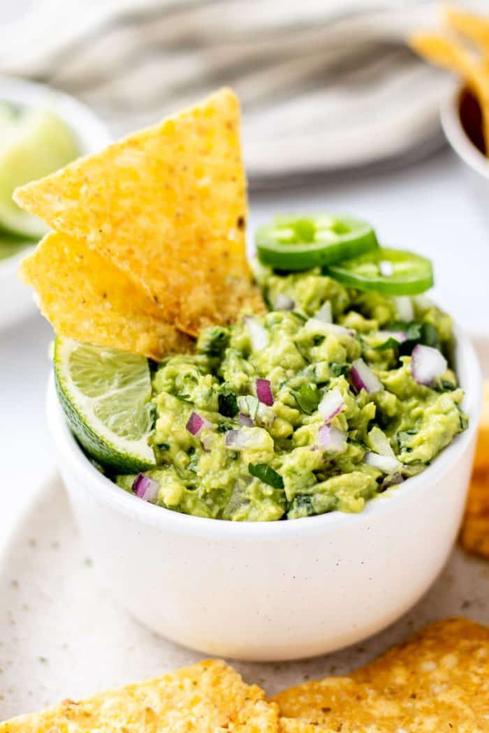 Chipotle Guacamole Dip in a white bowl garnished with a lime wedge, sliced fresh jalapeno, and tortilla chips. 