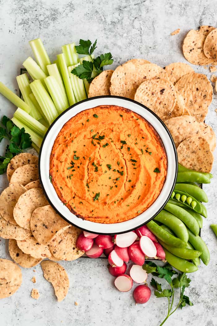 A White bowl surrounded by sliced veggie dippers and tortilla chips. In the bowl is an orange Buffalo Cauliflower Dip. 