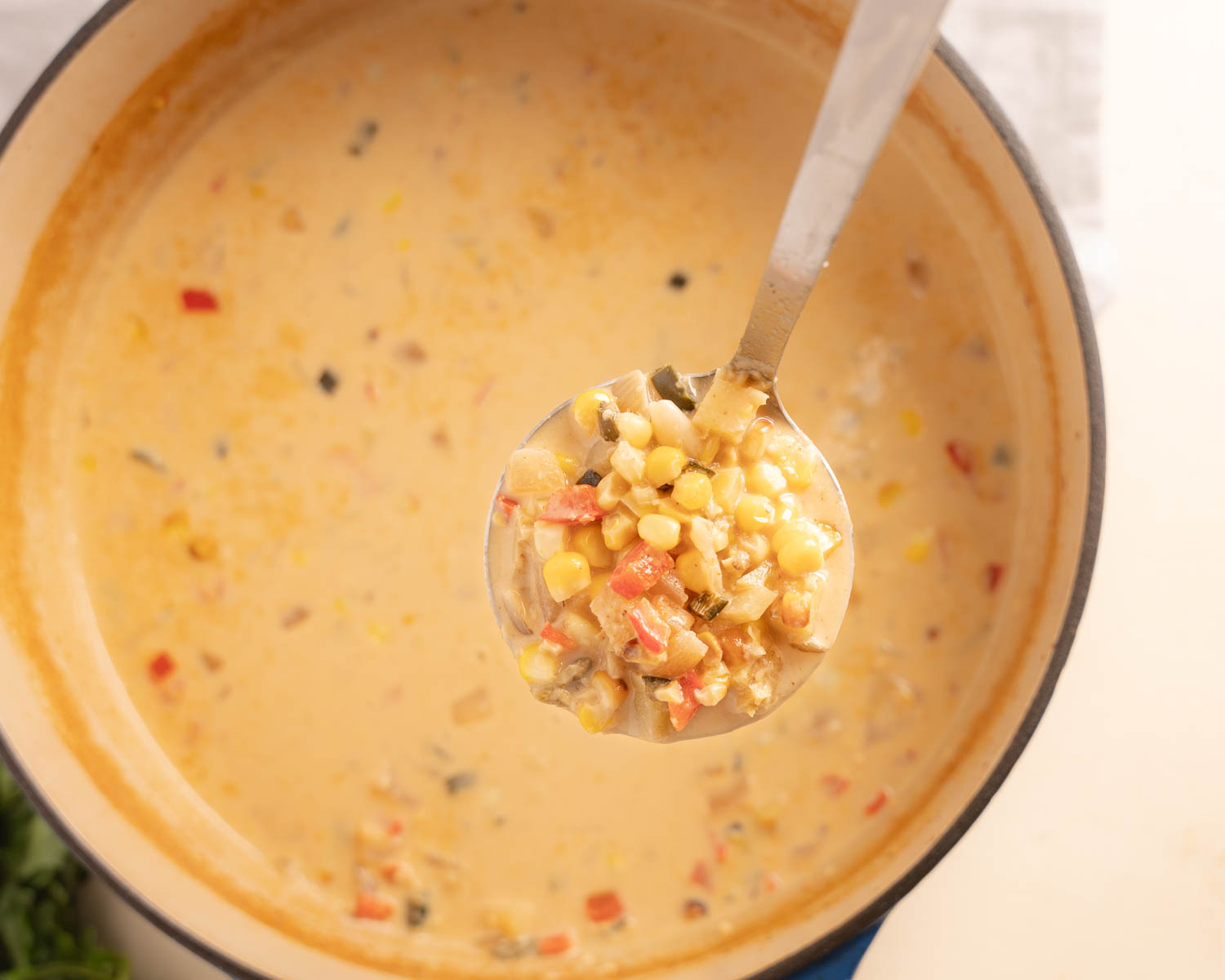 Top down of a dutch oven filled with Mexican Street Corn Soup, a laddle is hovering over the pot and it is filled with corn, red pepper speckles and soup. 