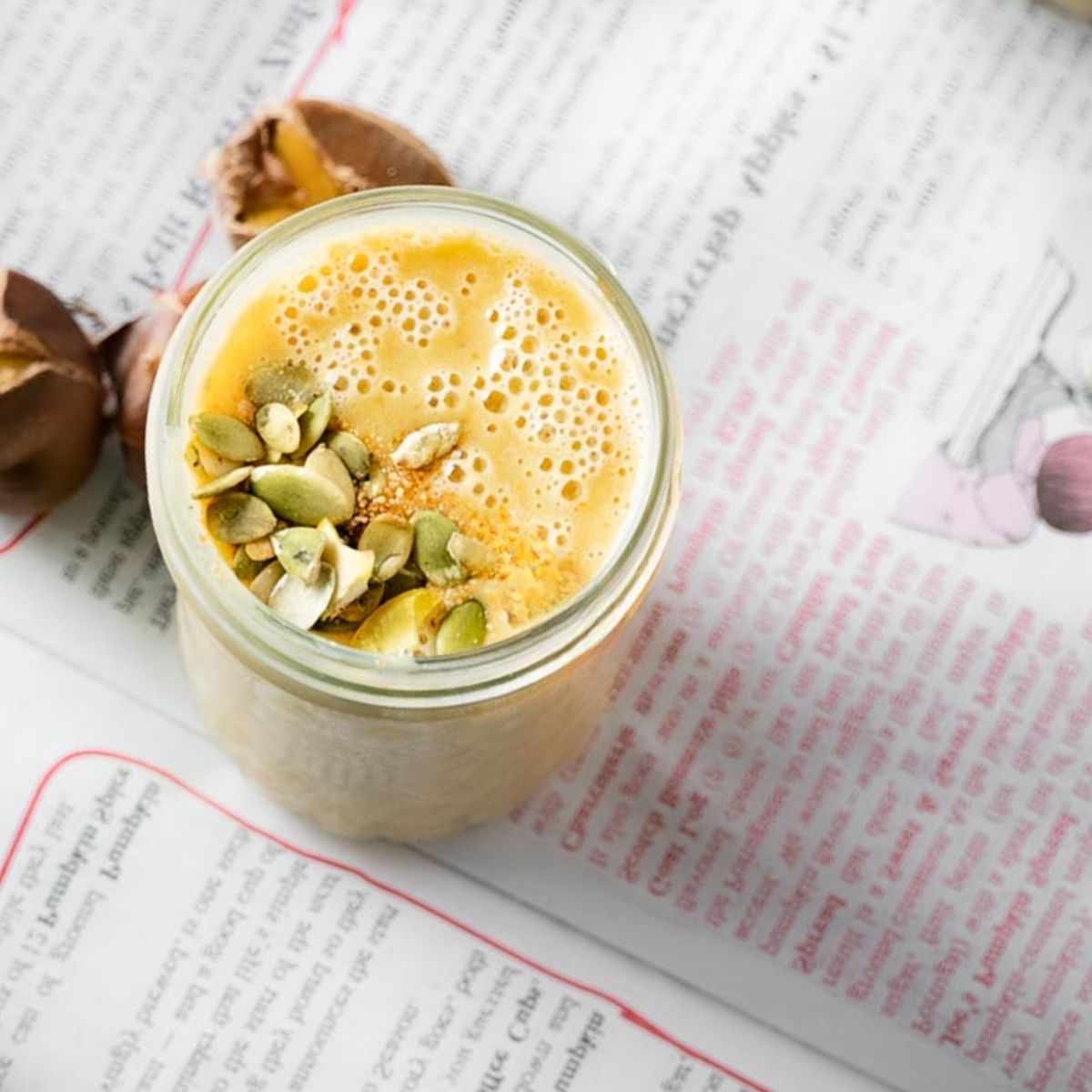 An 8oz mason jar filled with light orange pumpkin smoothie with chestnuts over newspaper with chestnuts scattered on the newspaper